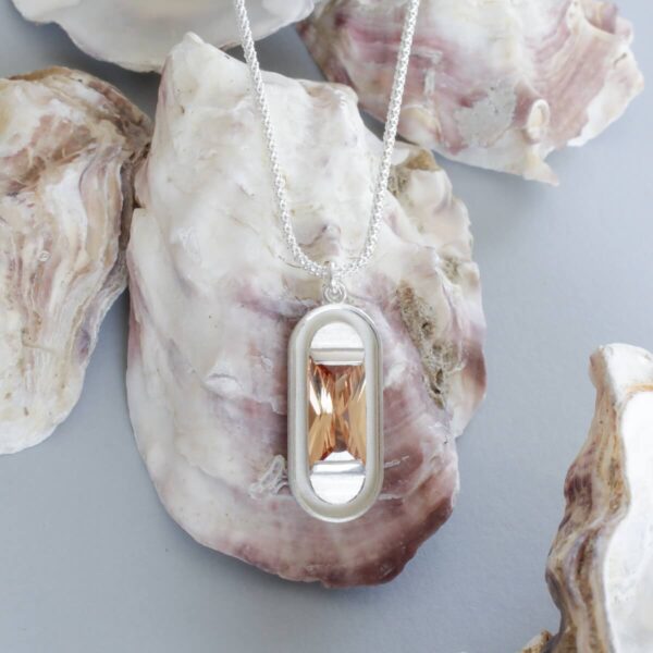 Hõbedast kaelakee modernse ristkülikukujulise ripatsiga, mille keskmes on hele šampanja toonis kivi. Ehe on pildistatud looduslikul merekarbi taustal, tuues esile selle pehme värvigamma ja orgaanilise esteetika. Minimalistlik disain ühendab kaasaegse vormi ja loodusest inspireeritud materjalitunnetuse, luues ajatult elegantse ehte igapäevaseks kandmiseks või kingituseks.